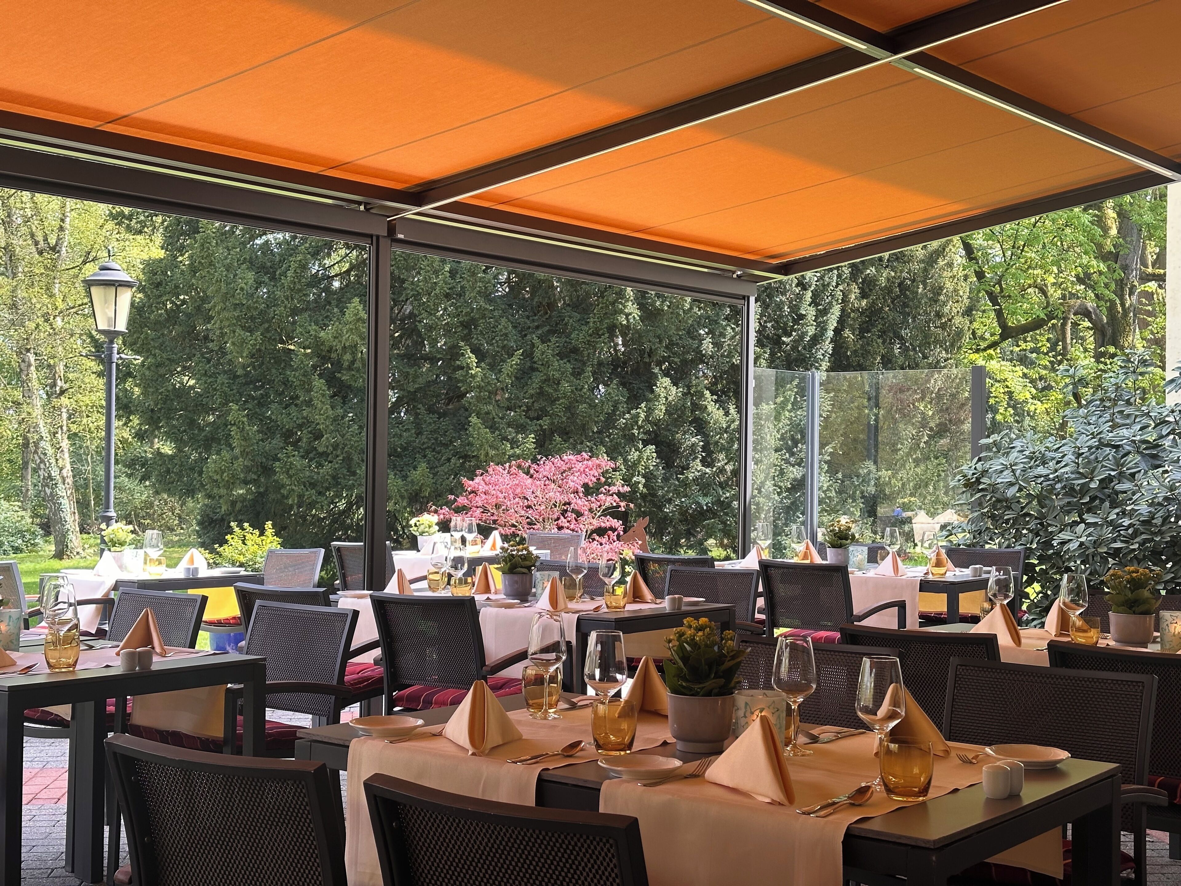 An outdoor dining area with tables set for two, covered by a canopy, and surrounded by lush greenery.