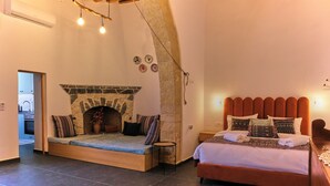 A bedroom with a stone fireplace, a bed with patterned pillows, and a view into the kitchen.