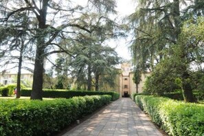Un chemin pavé, bordé de haies et d’arbres soigneusement taillés, mène à un bâtiment à la façade symétrique.