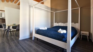 Una cama con dosel y cuatro columnas, con una colcha azul oscuro, una mesita de noche pequeña y una silla en una habitación moderna con vigas de madera y una kitchenette al fondo.