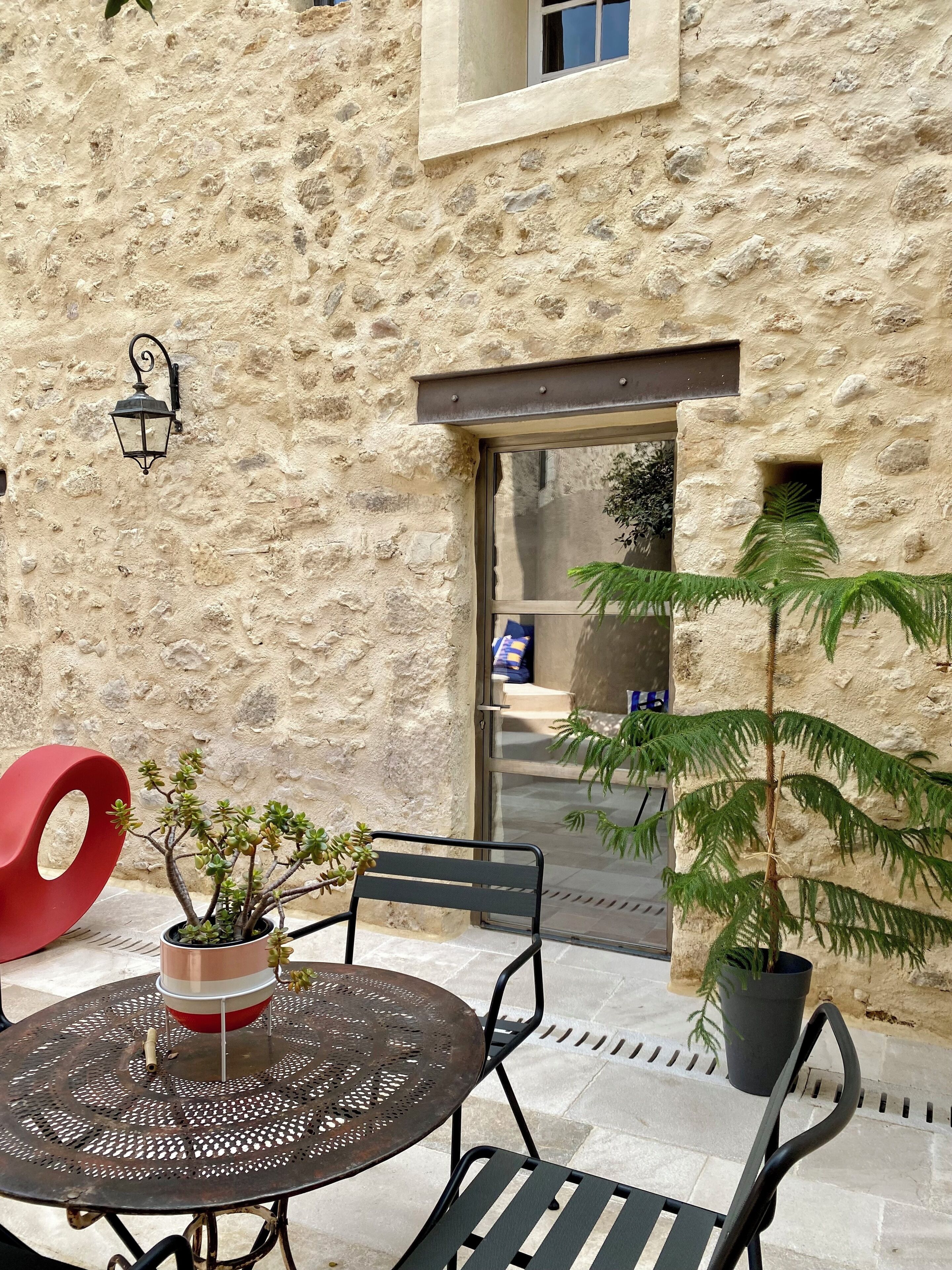 Un área de estar al aire libre con una mesa redonda de metal y dos sillas, un objeto circular rojo y plantas en macetas.
