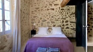 A bedroom with a stone wall, a bed with a purple bedspread, a bench, a nightstand, and a window with curtains.