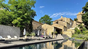 A swimming pool with lounge chairs and umbrellas, stone buildings, and trees in the background.
