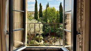 Finestra oberta amb vista a un jardí, una cadira de fusta i una barana de metall.