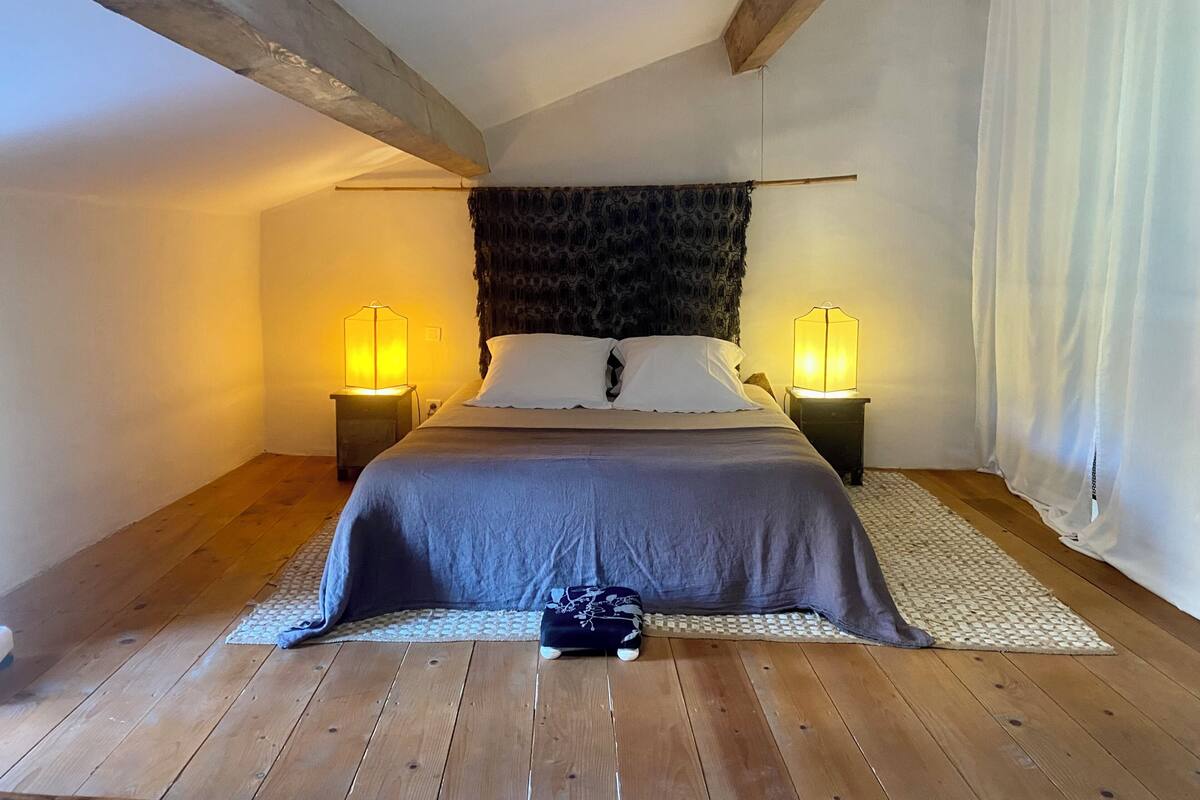 Ein Schlafzimmer mit einem Bett, zwei Nachttischlampen, einem dunkel gemusterten Kopfteil, Holzbalken und einem gewebten Teppich.