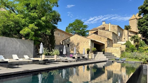 Una piscina amb gandules i ombrel·les, edificis de pedra i cel serena.