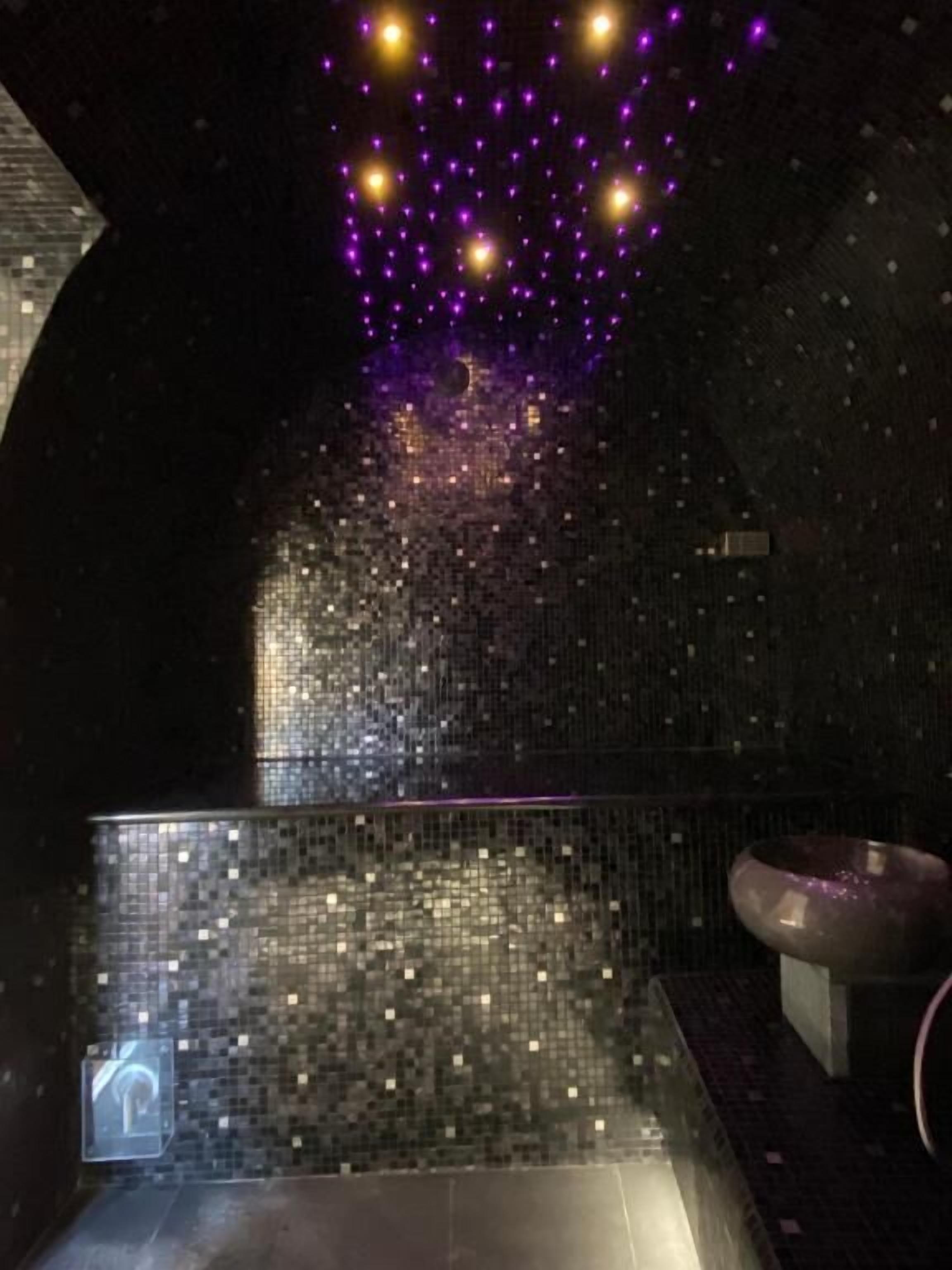 A bathroom with a mosaic wall, a sink, and a ceiling with purple lights.