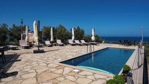 A poolside area with lounge chairs and umbrellas, a stone patio, and a view of the sea. - Thea Luxury Residences (Zacharo)