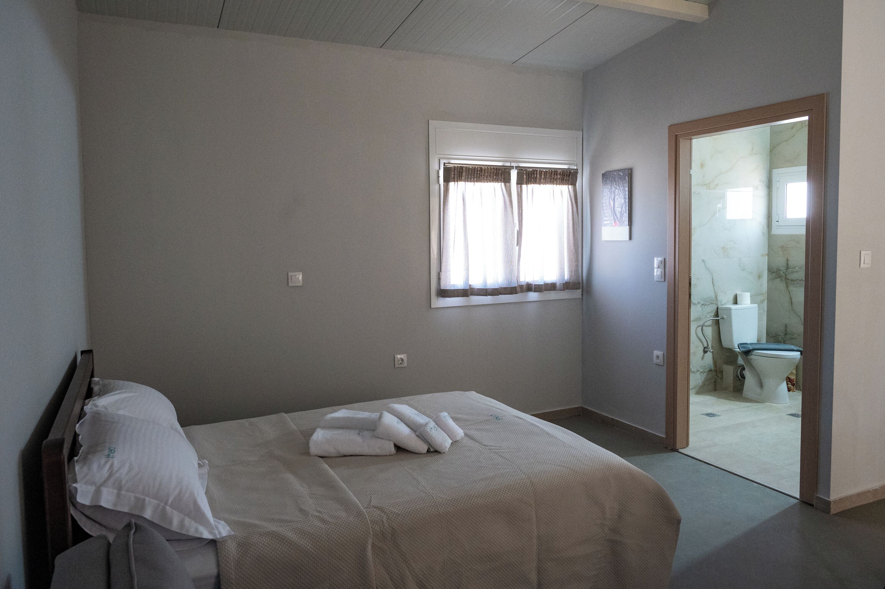 A bedroom with a bed, a window with curtains, a door leading to a bathroom, and a wall-mounted light switch.