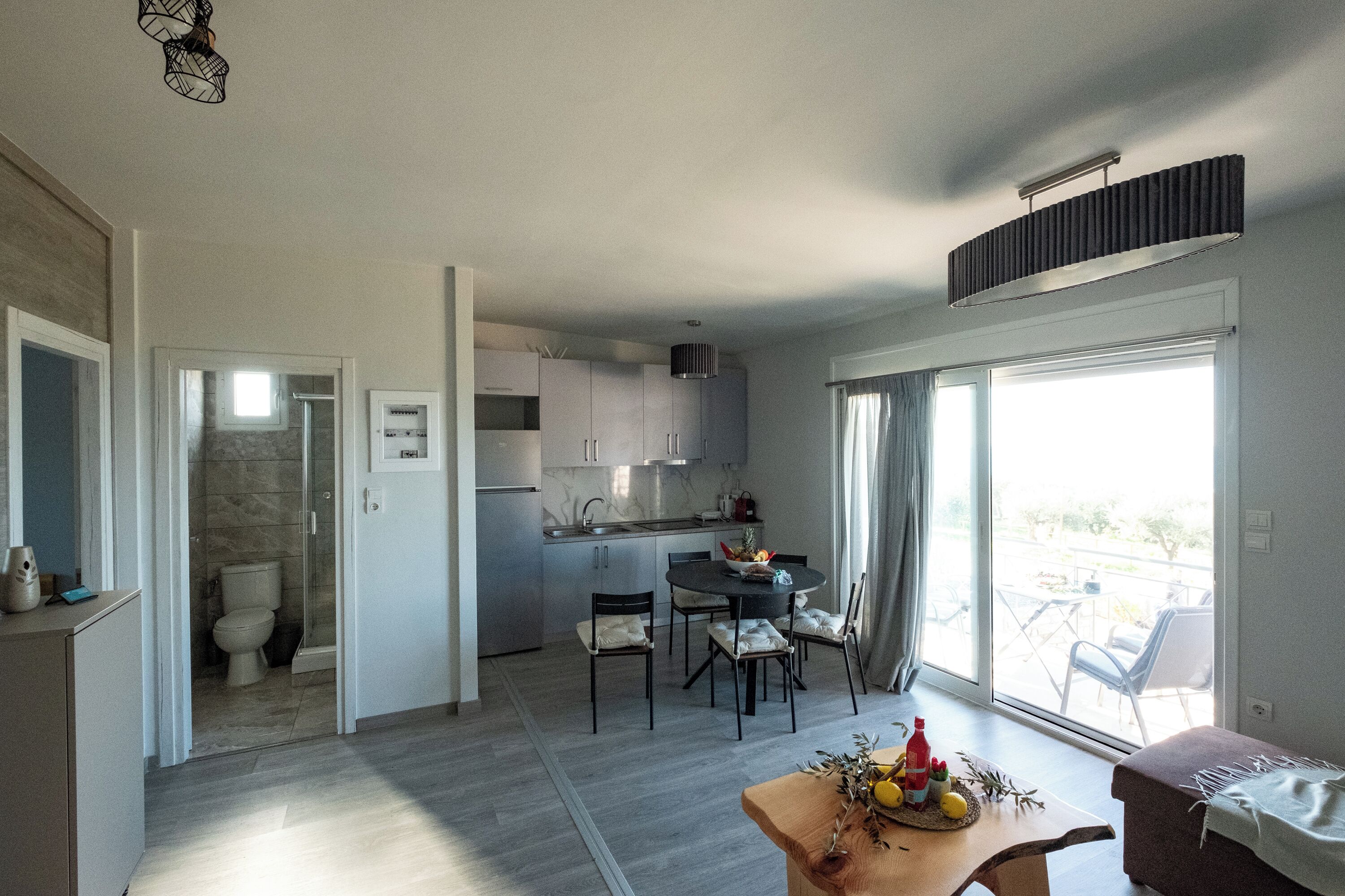 A modern kitchen with a dining area, a small table with fruit, and a view through the glass door.