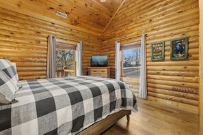 Ein Blockhauszimmer mit einem Bett, einem Fernseher und zwei Fenstern mit Vorhängen.