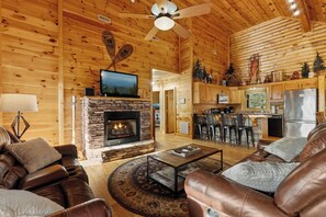 A log cabin interior with a stone fireplace, leather sofas, a dining area, and a kitchen.