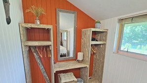 Een kamer met een houten wastafelmeubel, een spiegel, een klein bankje en een raam met jaloezieën.