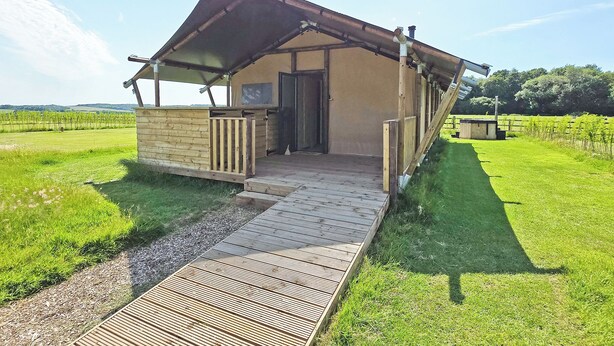 Een klein, laag gebouw met een schuin dak, een houten terras dat naar de ingang leidt, en een grasveld op de achtergrond.