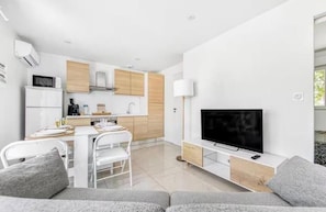 Uma sala de estar moderna com televisão de tela plana, um sofá, um espaço de refeições com cadeiras e uma cozinha com armários de madeira e eletrodomésticos brancos.