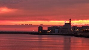 A coastal cityscape at sunset with a prominent tower structure and buildings along the waterfront. - VENTO DI BOLINA (Giovinazzo)