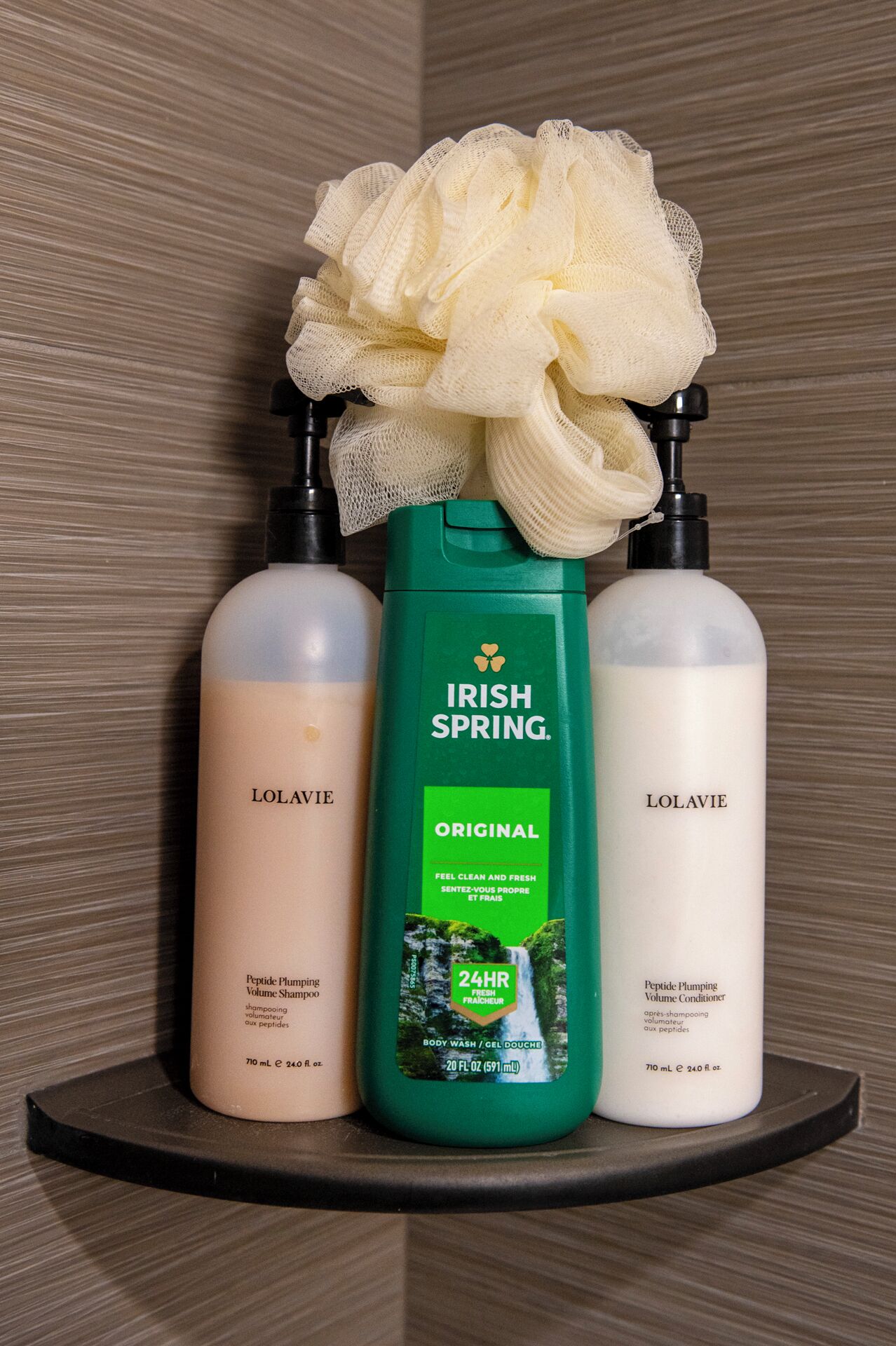 Three bottles of L'olavie shampoo and conditioner on a shelf with a shower sponge.