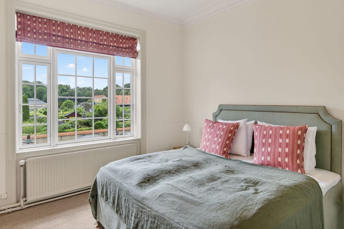 Une chambre avec un lit, une fenĂȘtre donnant sur un paysage verdoyant et des bĂątiments, et un radiateur.