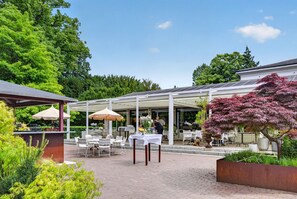 Un espace extérieur aménagé en terrasse, avec des tables et des chaises, un coin salon abrité et un bac à fleurs contenant un arbre.