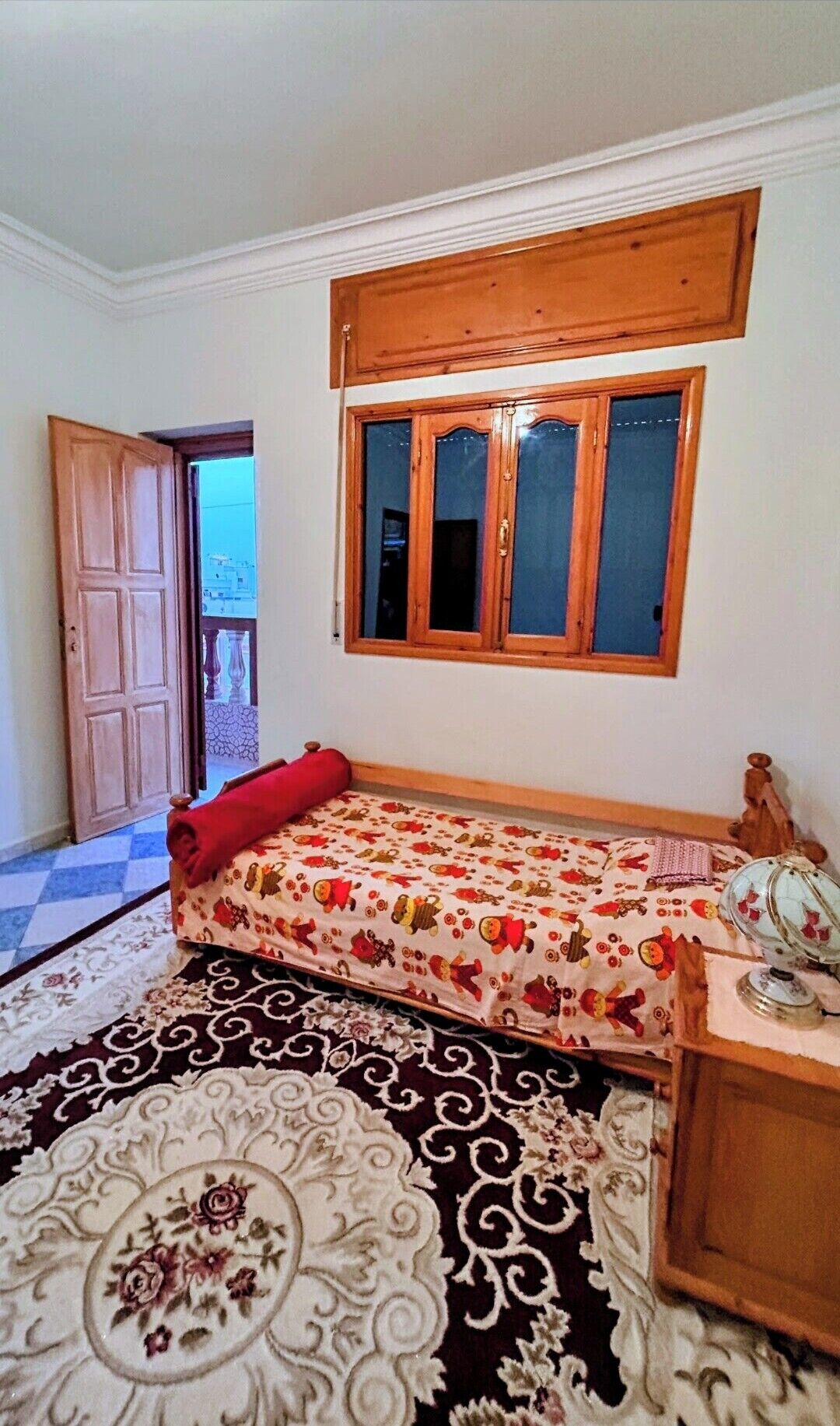 Een kamer met een bed, een raam, een deur en een kamerbreed tapijt met dessin.