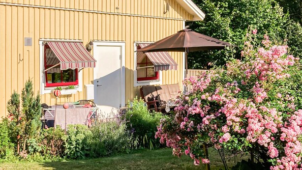 En gul bygning med en dør, to vinduer med markiser, en terrasse med bord og stole, og en blomstrende busk.