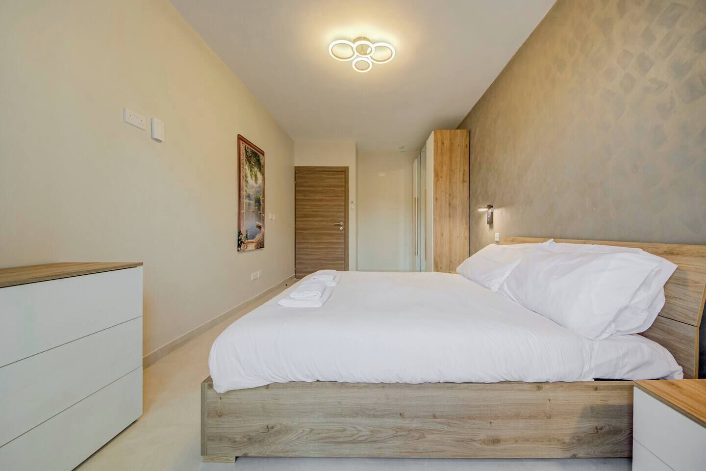 A bedroom with a wooden bed, a nightstand, a wall-mounted light, and a painting on the wall.