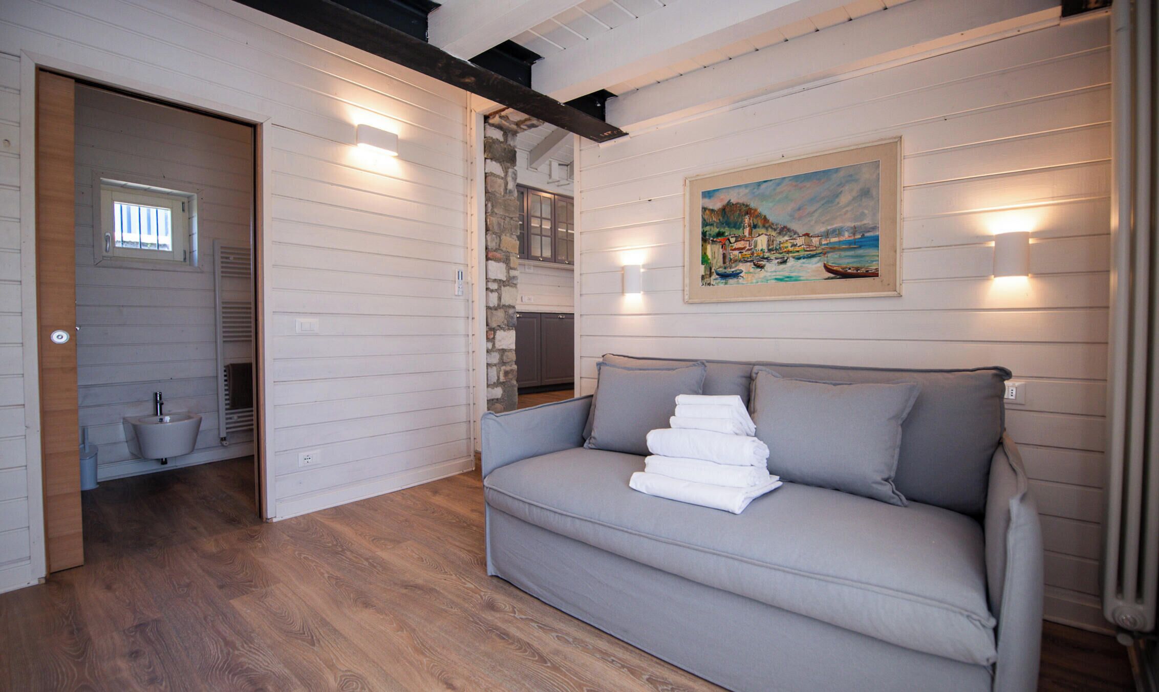 A room with a grey sofa, a painting of a coastal scene, and a stone wall.
