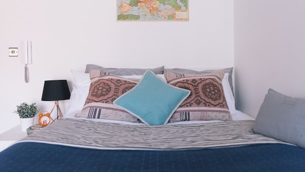Una cama bien hecha con almohadones estampados, un mapa colgado en la pared, una lámpara de noche y una pequeña planta.