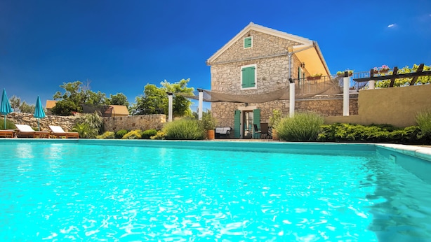 Una piscina con sillas de descanso, un edificio de piedra con una persiana verde y un cielo azul despejado.