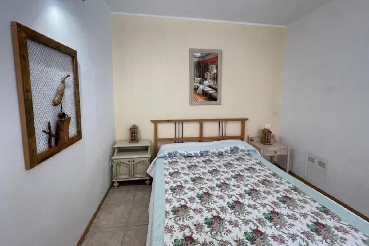 Un dormitorio con cama, mesita de noche, cómoda, un cuadro enmarcado en la pared y un mueble pequeño.