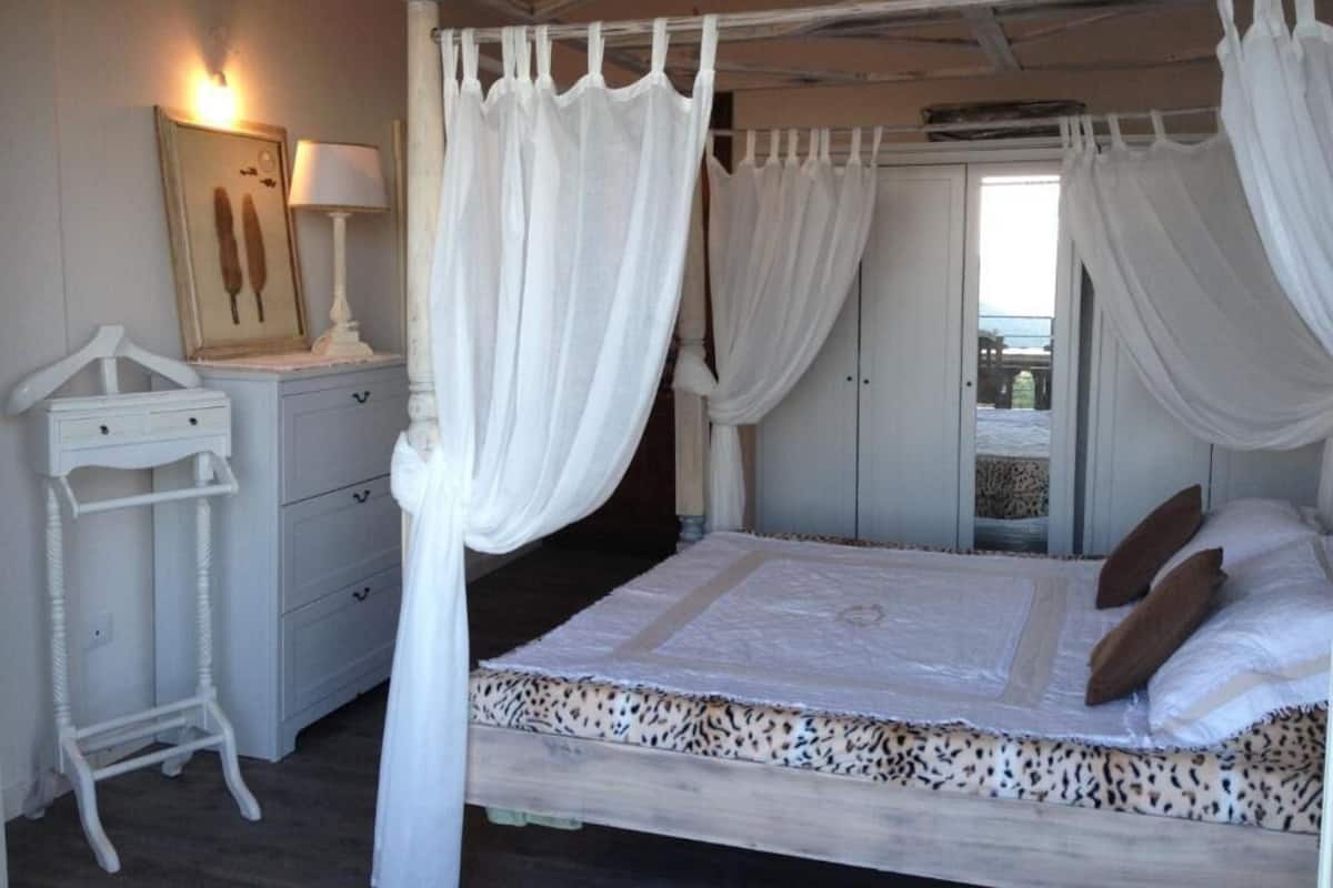 Una cama con dosel adornada con cortinas blancas, una mesita de noche con lámpara y un cuadro enmarcado en la pared.