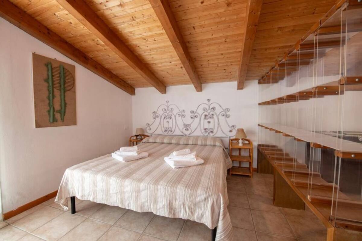 Un dormitorio con techo de madera, una cama con colcha a rayas, cabecera con diseño decorativo y una escalera con barandilla de cristal.