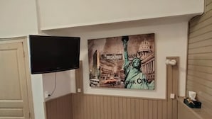 Een kamer met een aan de muur bevestigde televisie, een poster van de Vrijheidsbeeld en Broadway, en een houten paneelwand.