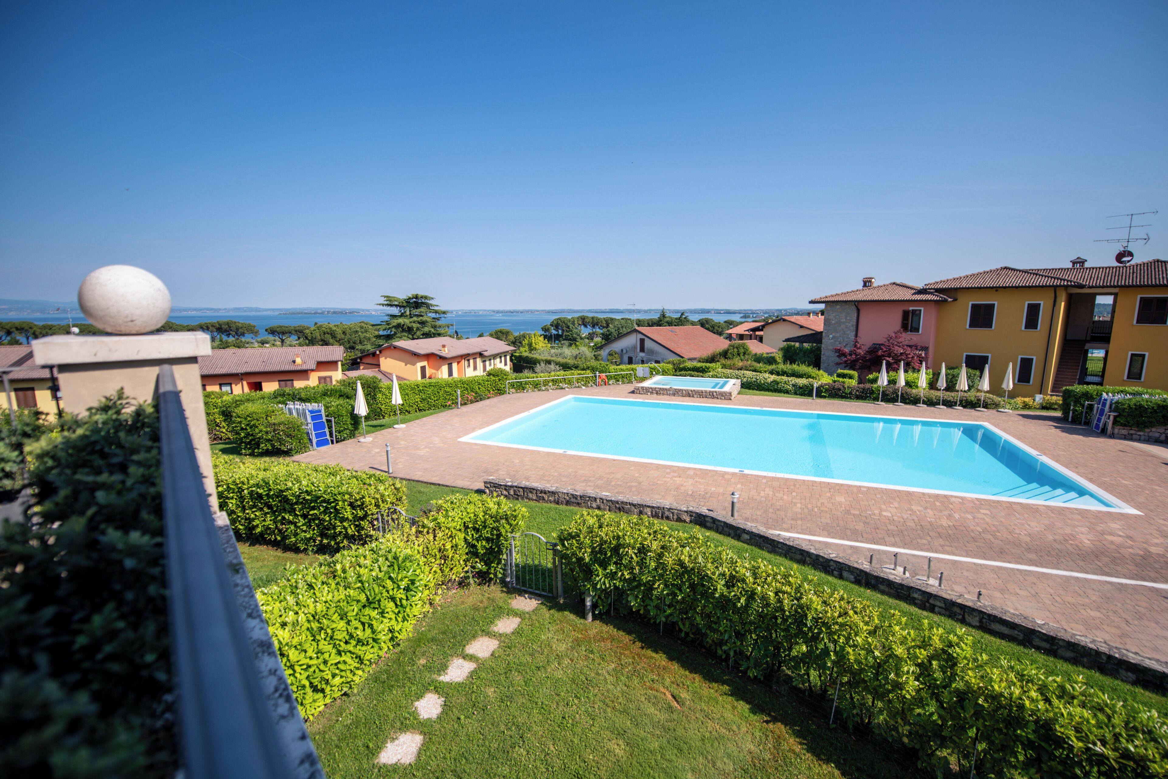 A swimming pool surrounded by greenery and buildings, with a clear view of the sea in the background.
