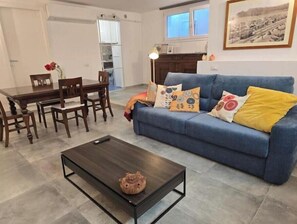 A living room with a blue sofa, a coffee table, and a dining area with a wooden table and chairs. - Cozy Studio by the Sea - Relaxation between beaches and pool in Sardinia (Quartu Sant'Elena)