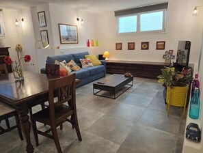A living room with a blue sofa, a dining table with chairs, a coffee table, and a window with blinds. - Cozy Studio by the Sea - Relaxation between beaches and pool in Sardinia (Quartu Sant'Elena)