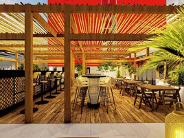 Un área de estar exterior de madera con mesas y sillas, un bar y una pérgola con tela a rayas rojas y amarillas.