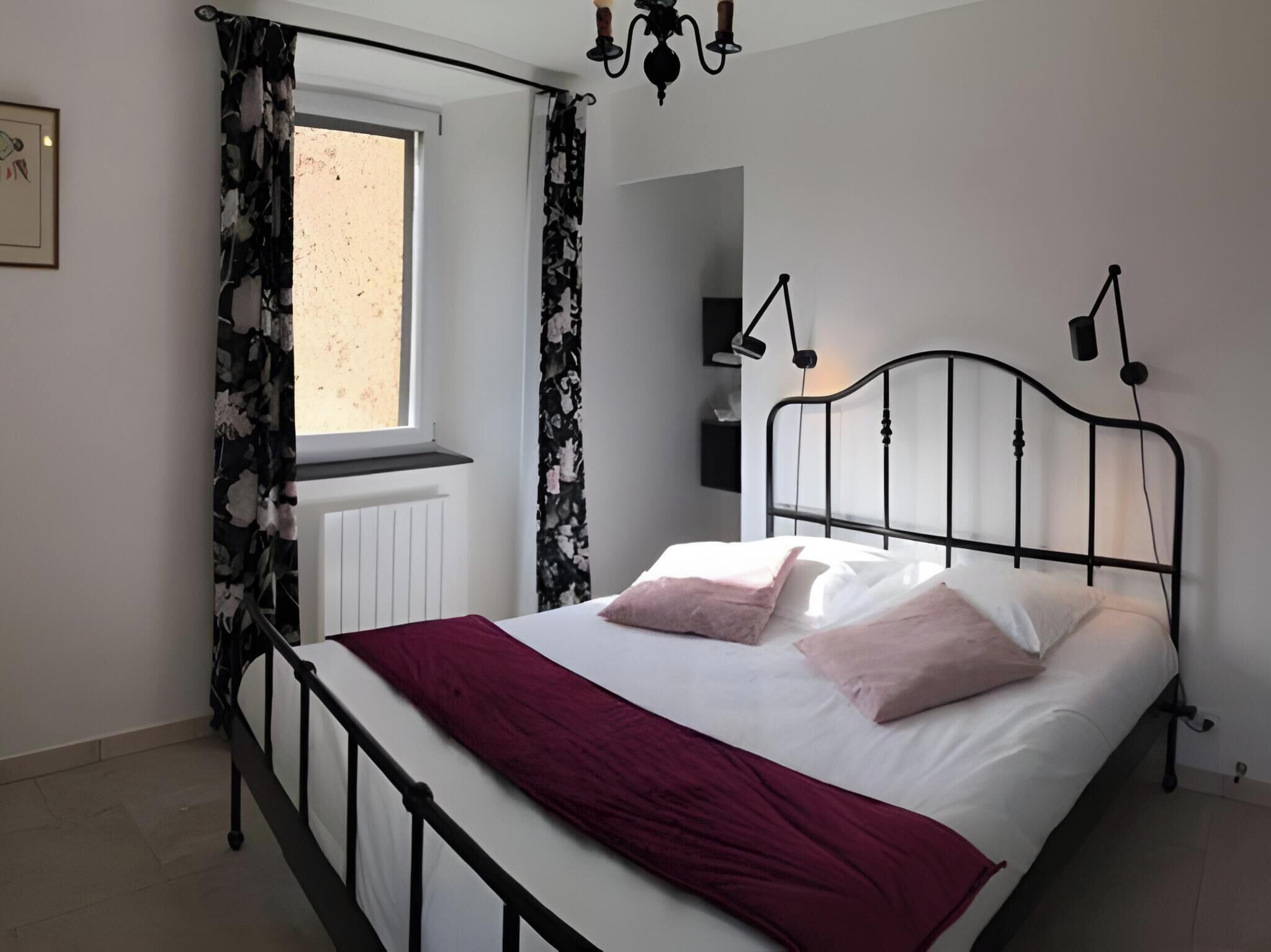 Ein Schlafzimmer mit Bett, einem Fenster mit Vorhängen, einem Heizkörper, einer Wandlampe und einem Wandfernseher.