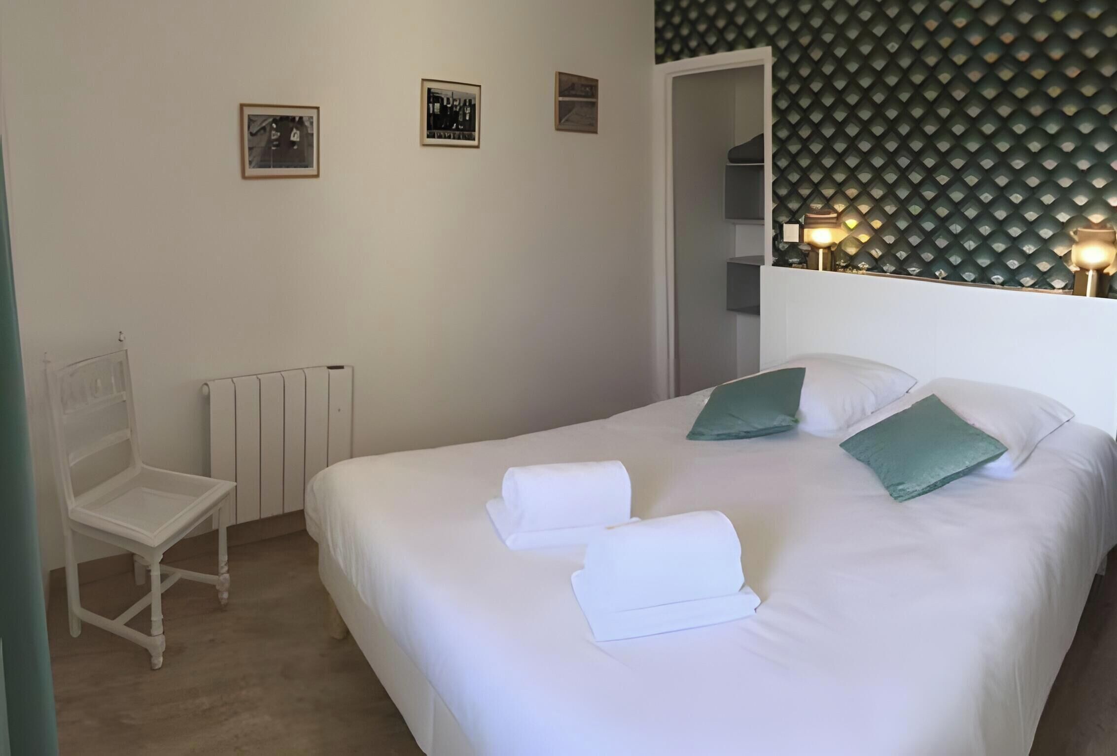 Una cama bien hecha con sábanas blancas y almohadas verdes, una silla blanca, un radiador y cuadros enmarcados en la pared.