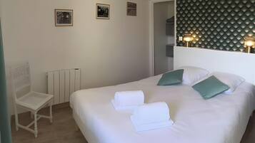 Una cama bien hecha con sábanas blancas y almohadas verdes, una silla blanca, un radiador y cuadros enmarcados en la pared.