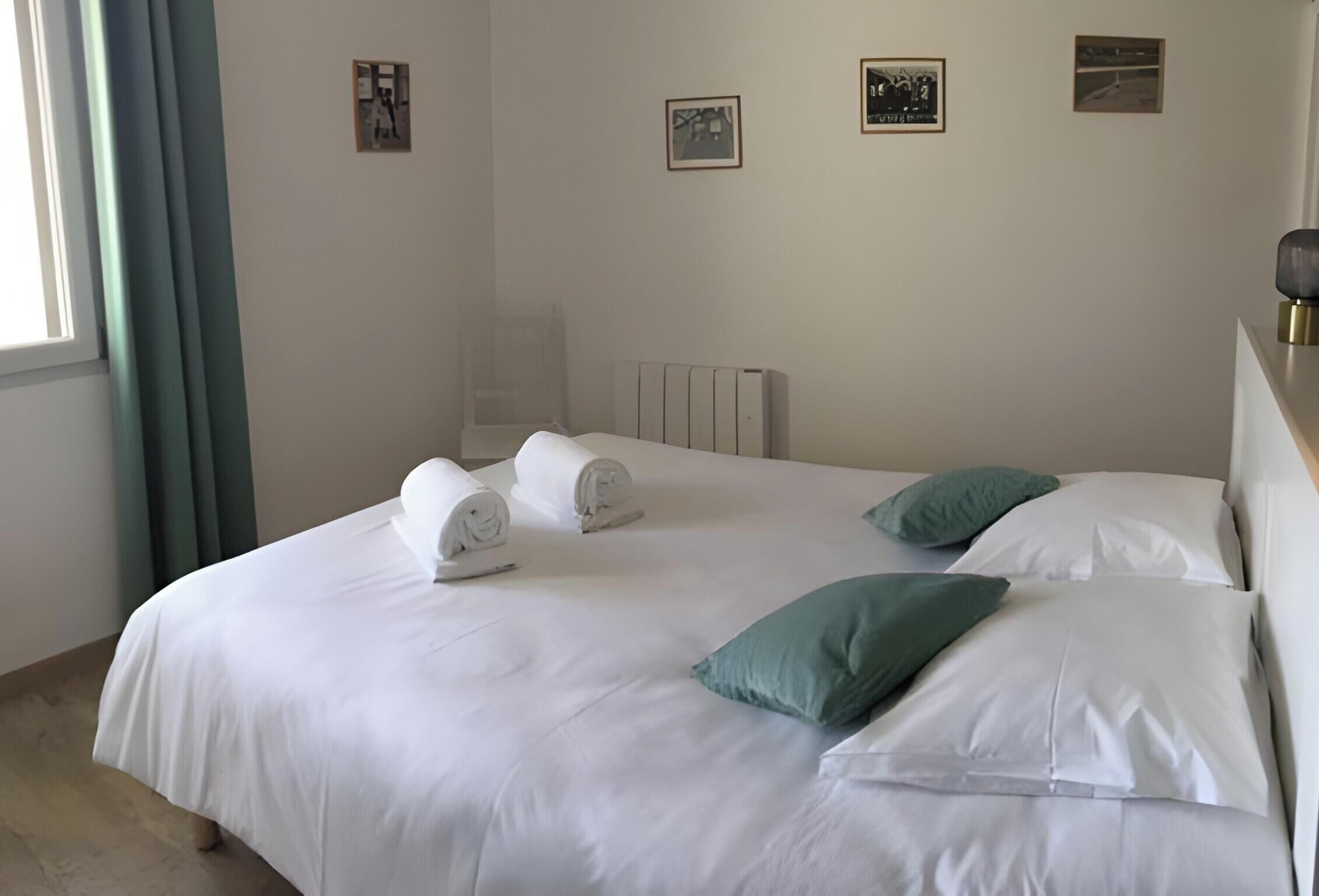 Een opgemaakt bed met witte lakens en twee groene kussens, een radiator aan de muur en een raam met gordijnen.