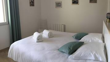 Een opgemaakt bed met witte lakens en twee groene kussens, een radiator aan de muur en een raam met gordijnen.