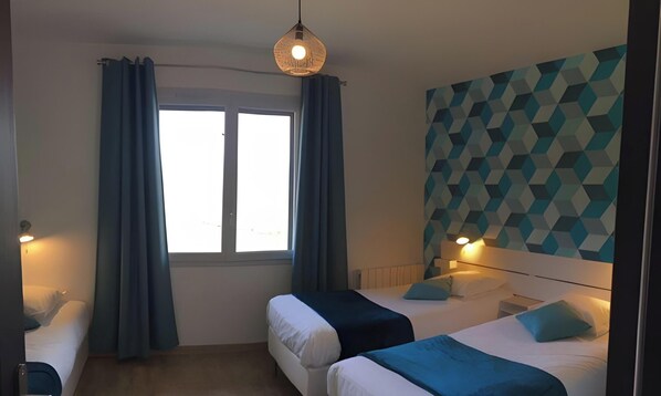 A room with two beds, a window with curtains, a geometric-patterned wall, and a hanging light fixture.