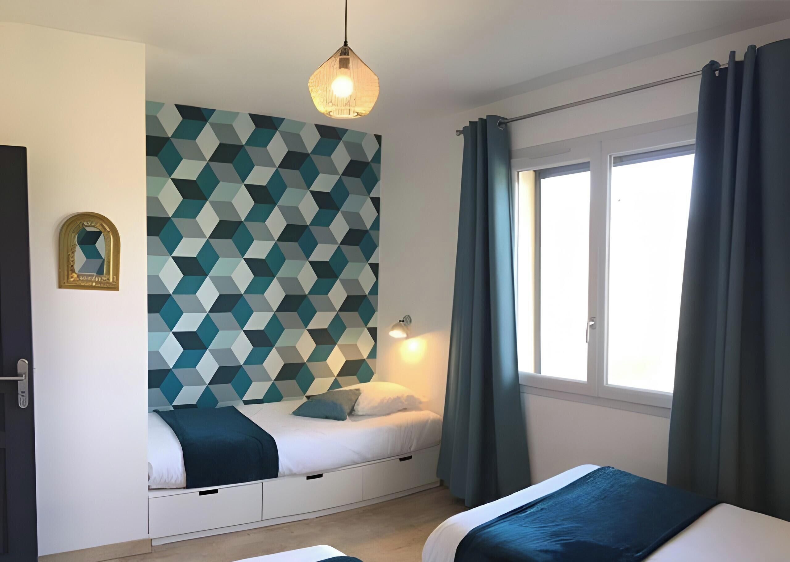 A bedroom with a geometric-patterned wall, a bed with a white headboard, a bedside table, a hanging light fixture, and a window with curtains.