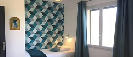 A bedroom with a geometric-patterned wall, a bed with a white headboard, a bedside table, a hanging light fixture, and a window with curtains.