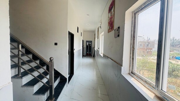 A corridor with a staircase on the left, a window on the right, and doors along the corridor. - Maa Vaishno Dham (Vrindavan)