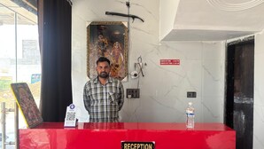 A reception desk with a 'RECEPTION' sign, a wall-mounted security camera, and a framed religious painting. - Maa Vaishno Dham (Vrindavan)