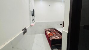 A small, clean room with white walls, a marble floor, a bed with a patterned bedspread, a ceiling fan, and a mounted television. - Maa Vaishno Dham (Vrindavan)