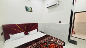 A room with a bed, a wall-mounted air conditioner, a painting, and a door leading to a bathroom. - Maa Vaishno Dham (Vrindavan)