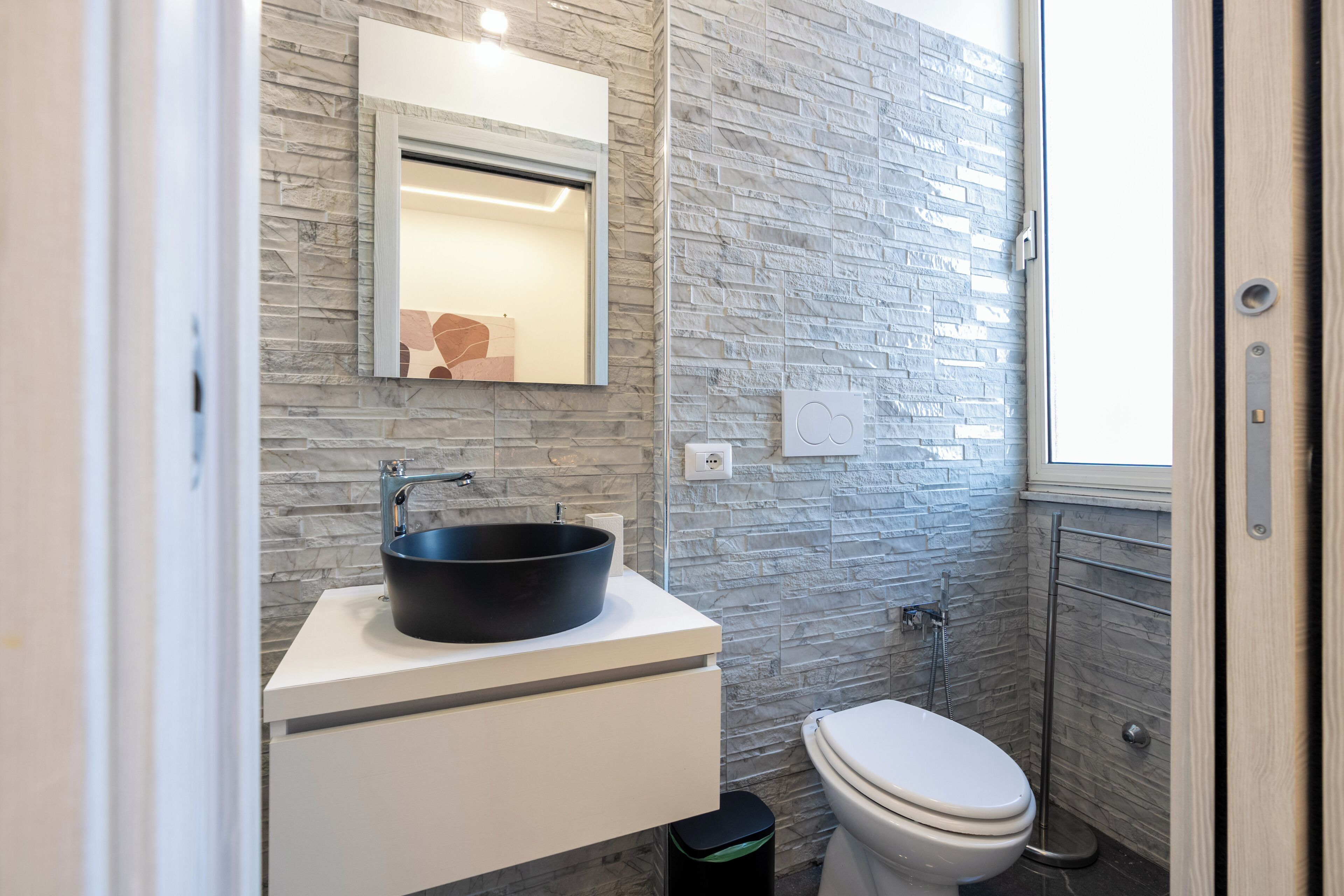 A bathroom with a stone wall, a black sink, a white toilet, and a mirror above the sink.